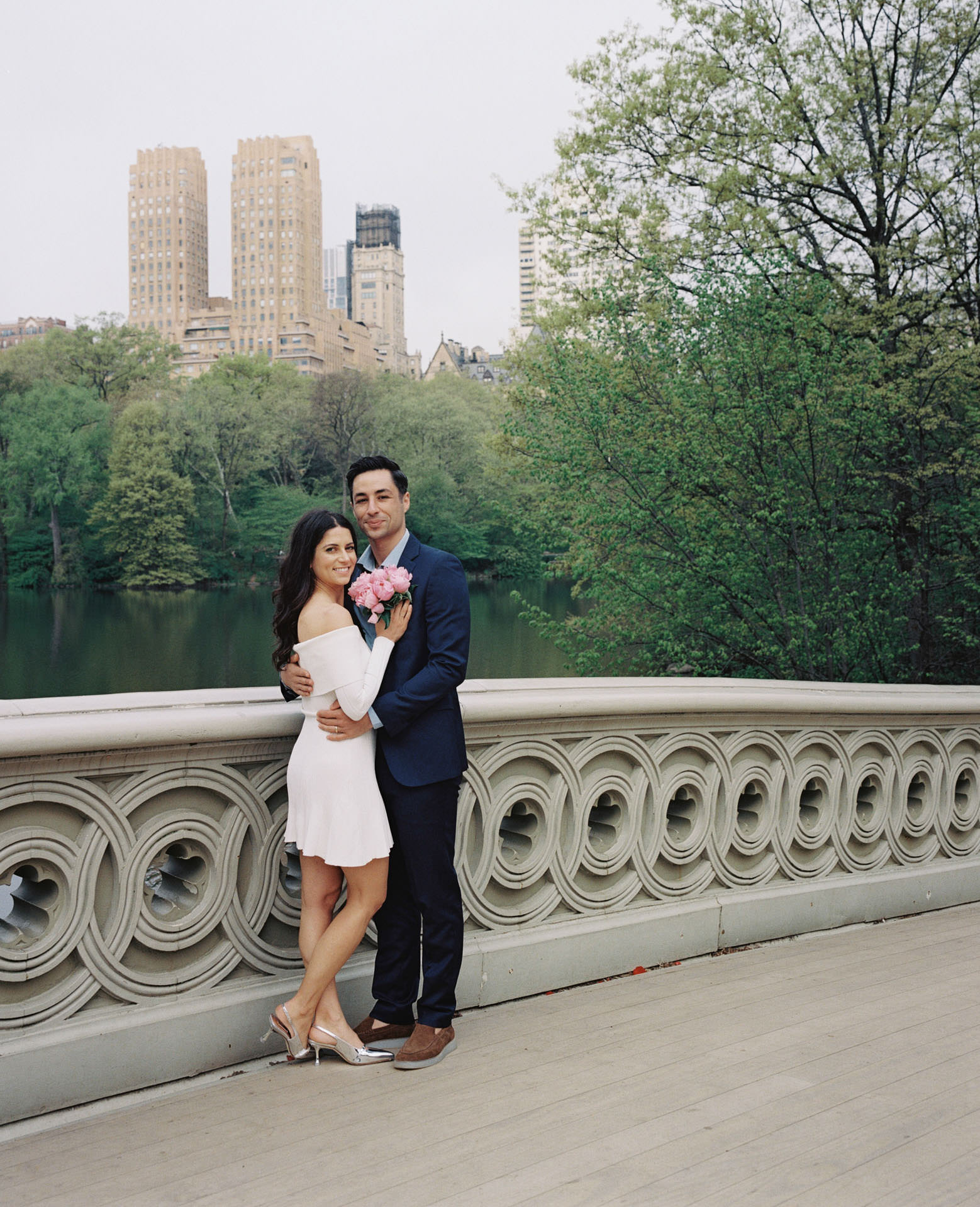 An engaged couple standing on Bow Bridge, with the picturesque New York City skyline in the distance.