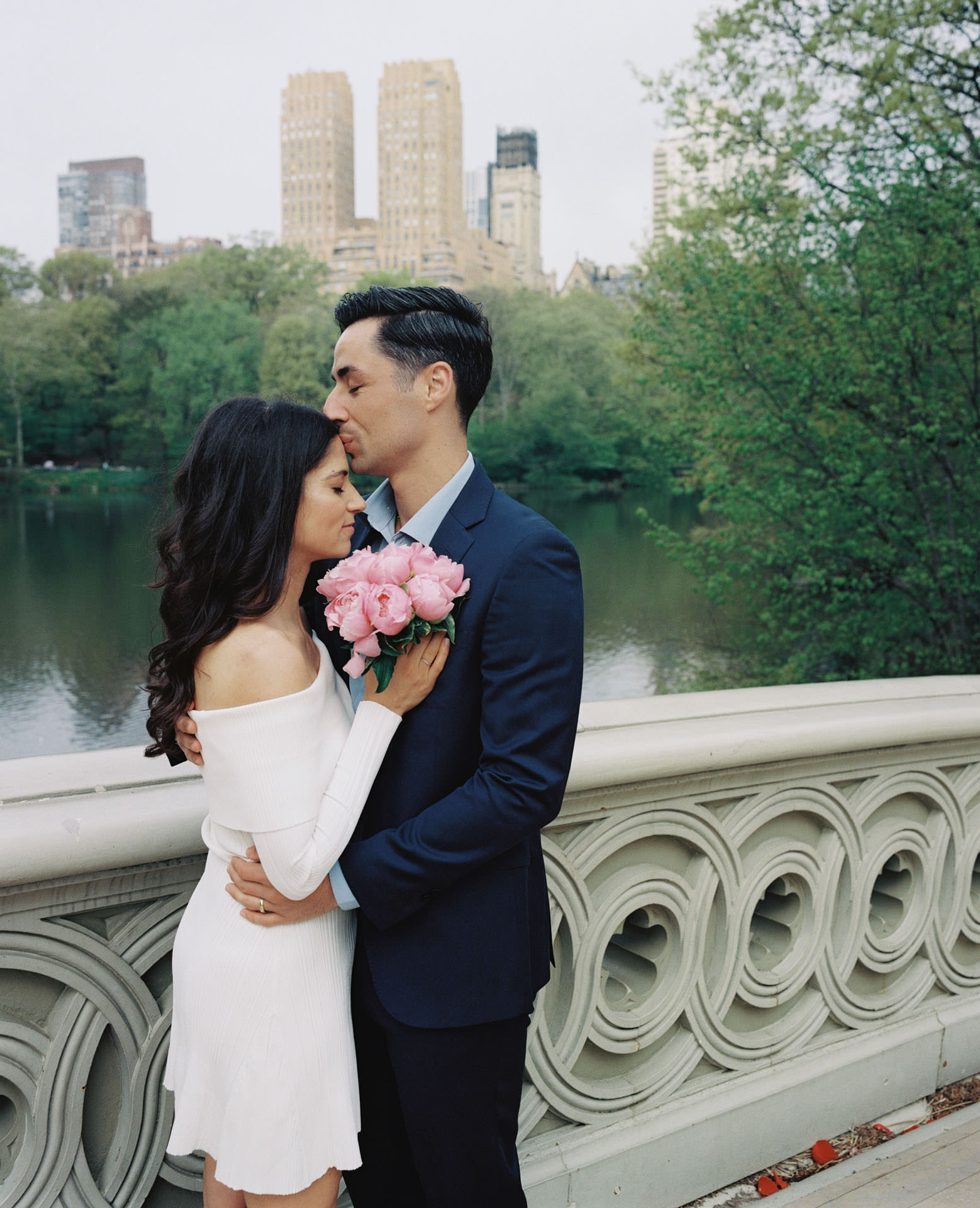 A couple sharing a kiss on the elegant Bow Bridge, with the serene lake and lush greenery in the background.