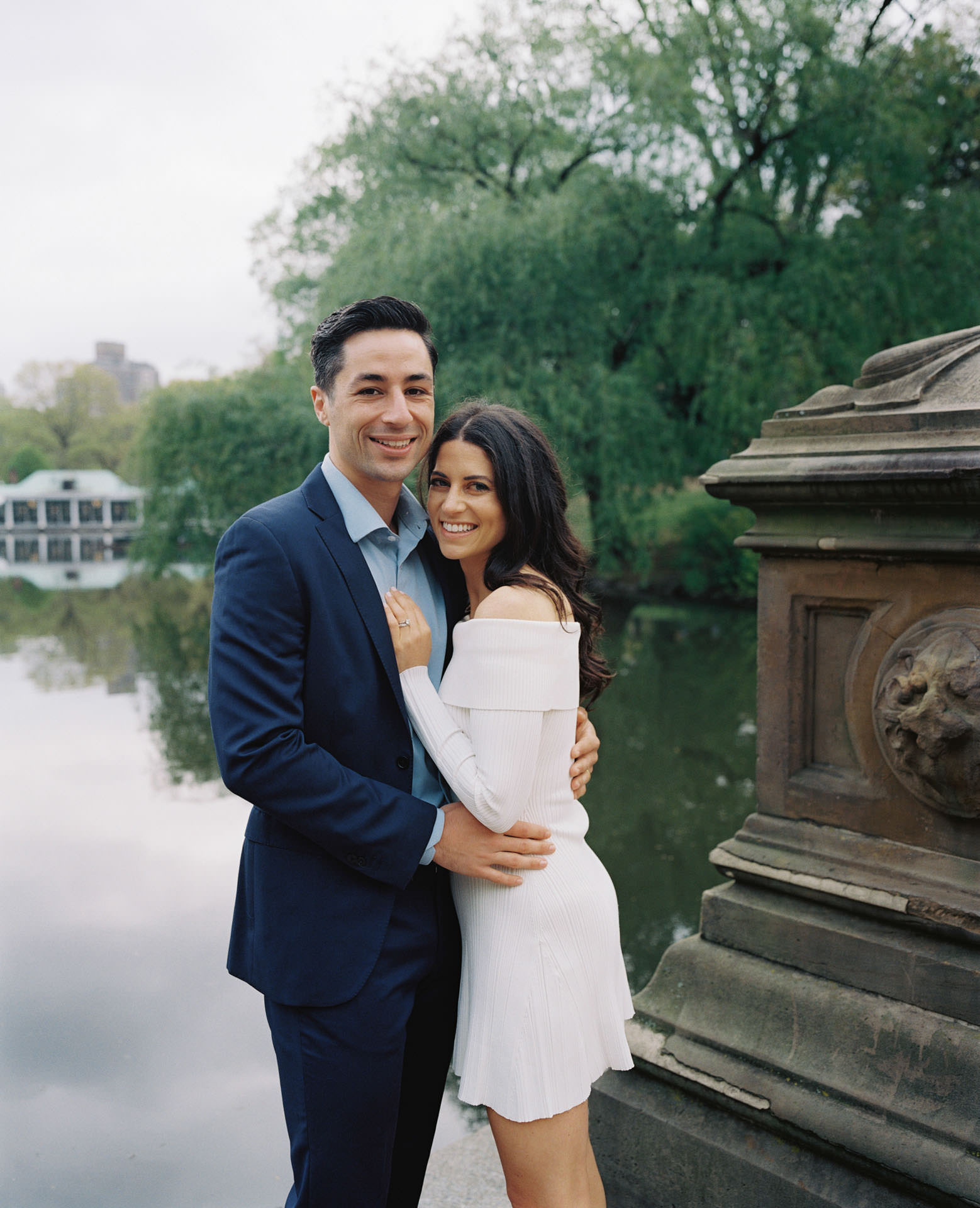 An engaged couple walking hand-in-hand along the water's edge near the Central Park Boathouse, with the charming boathouse in the background.