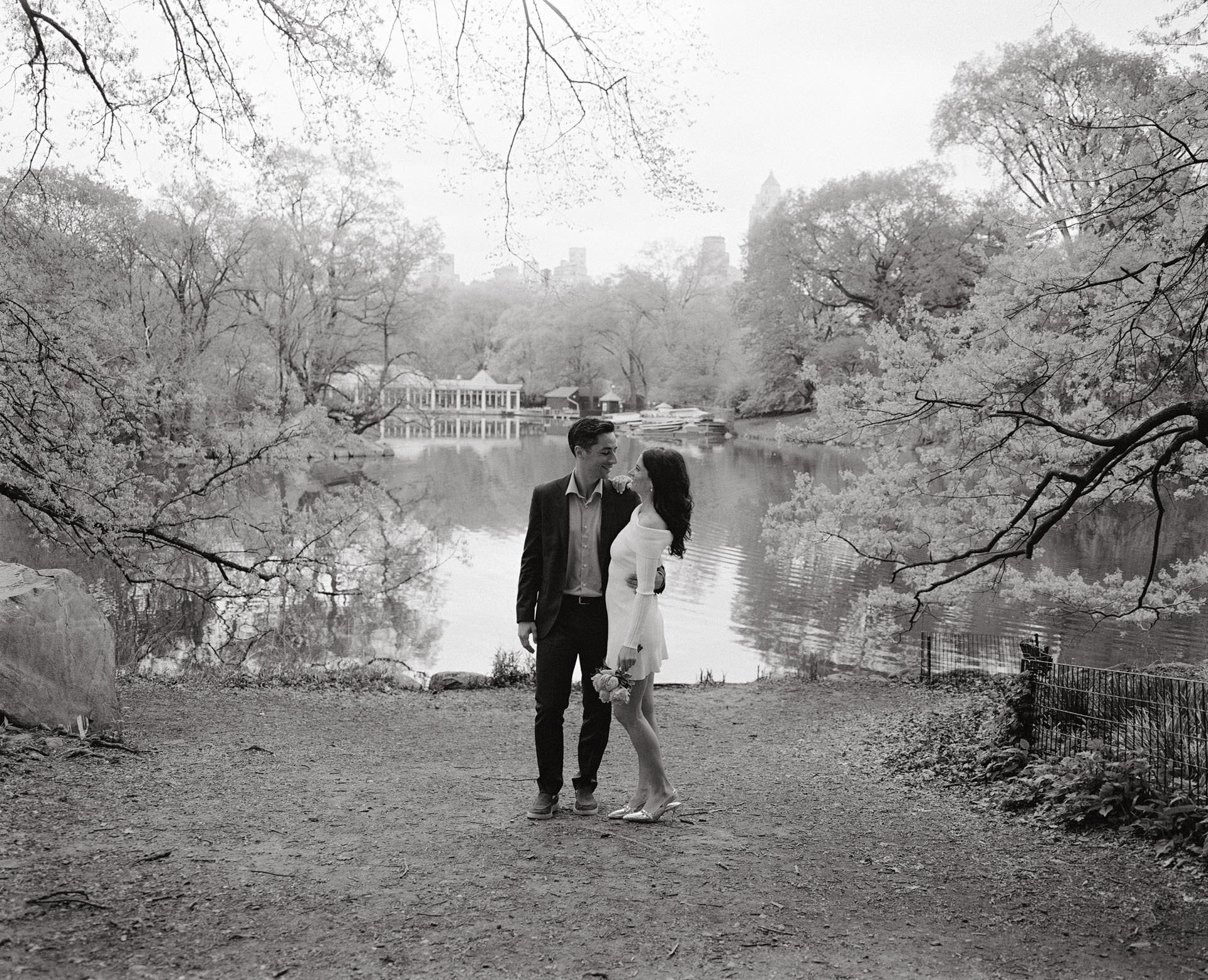 A couple enjoying a romantic walk next to the lake near the Central Park Boathouse, with the lush greenery reflected in the water.