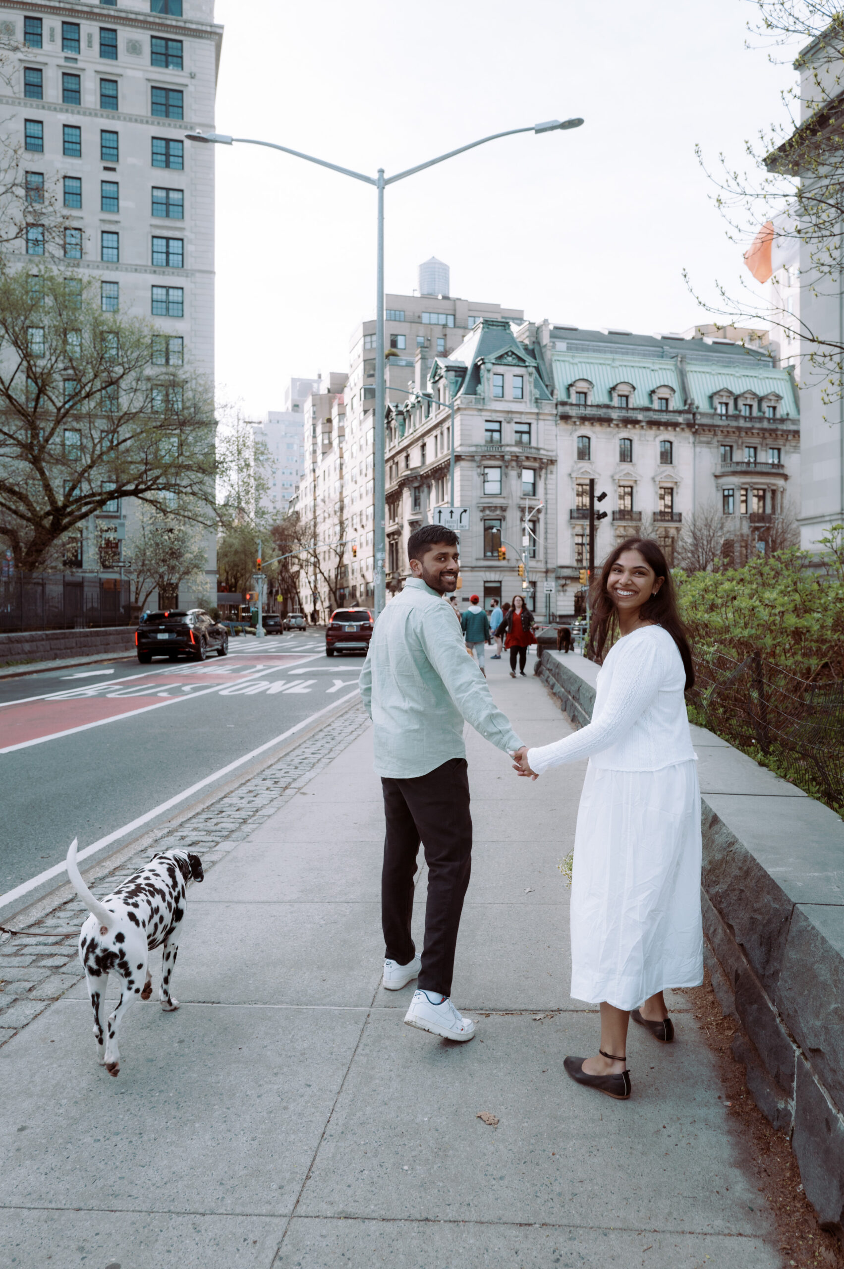 The engaged couple is walking hand in hand in a NYC sidewalk, with beautiful buildings in the background.
