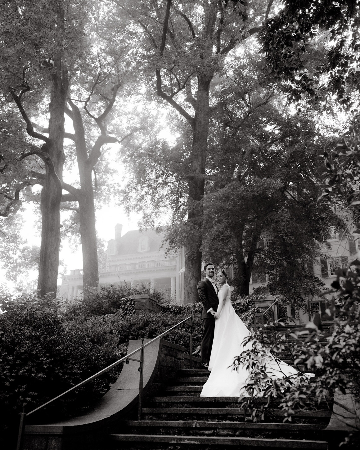 The elegant bride and groom are standing on a staircase in a garden with magnificent trees in the background taken by Jenny Fu Studio.