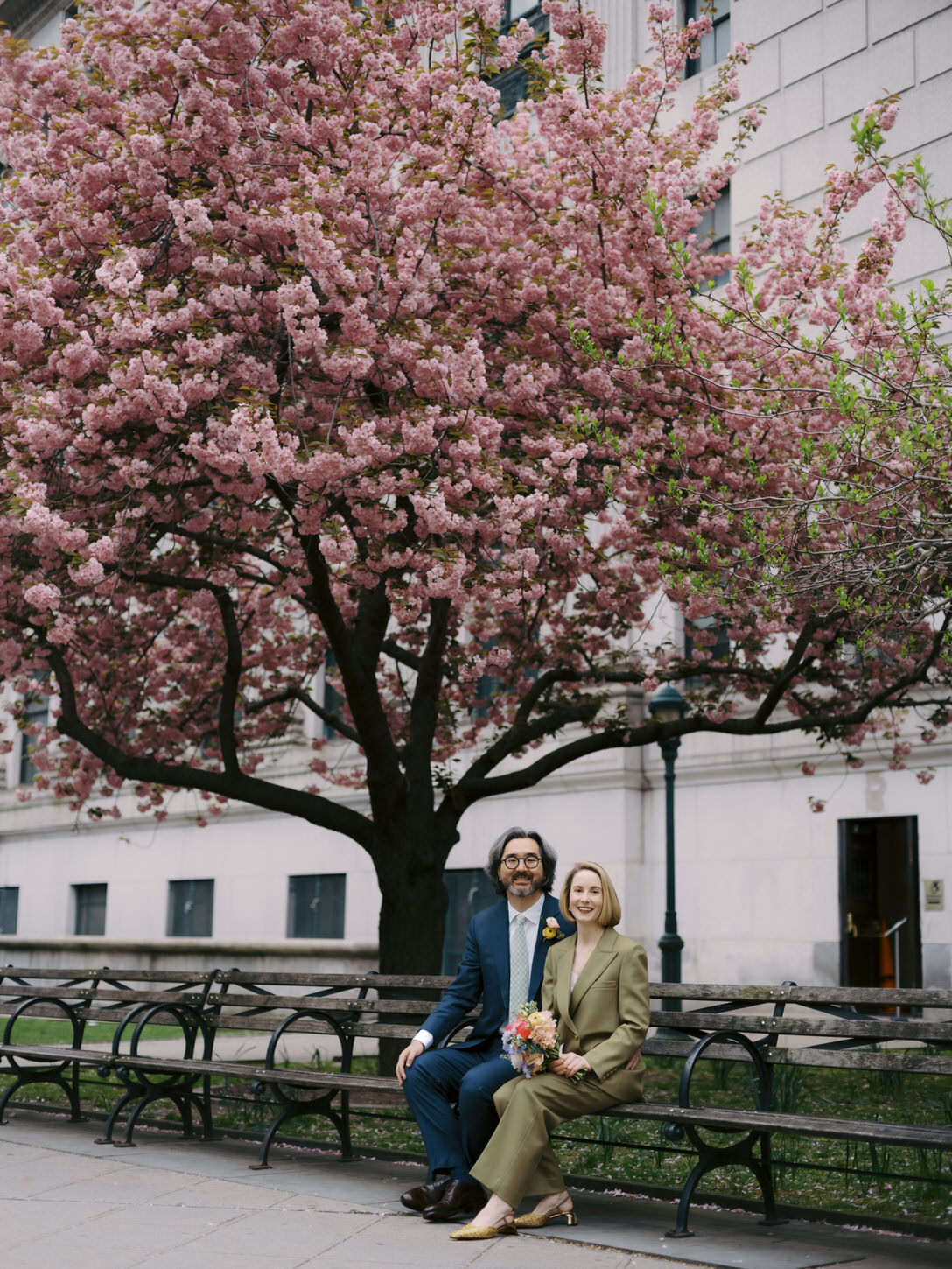 The newlyweds are sitting on a bench with cherry blossom trees in the background shot by Jenny Fu Studio, NYC.