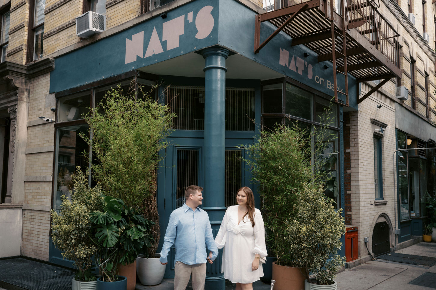 The sweet engaged couple are holding hands, smiling in front of Nat's on Bank restaurant in NYC, captured by Jenny Fu Studio.