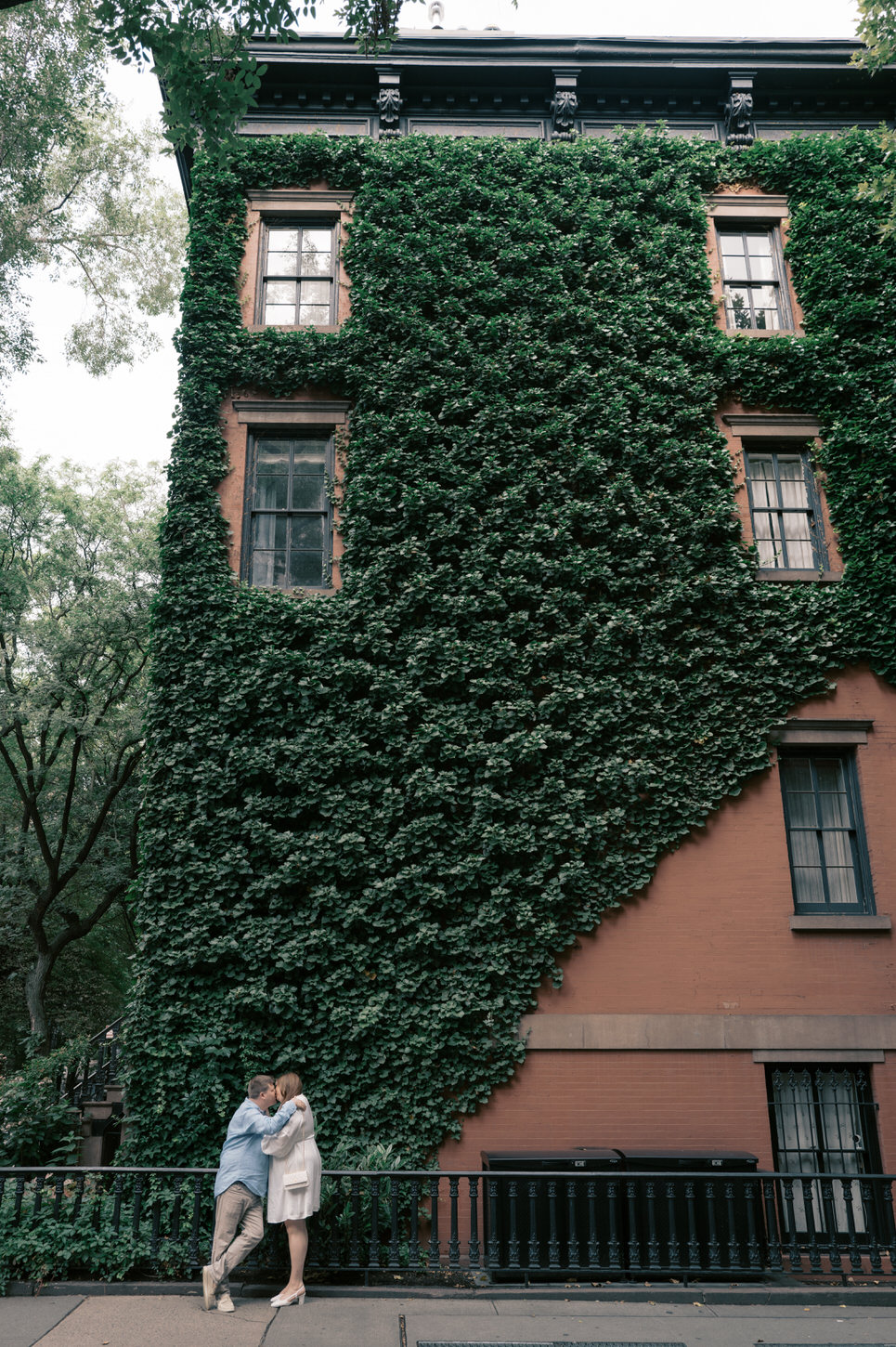 An engaged couple kissing each other with a beautiful ivy-covered building on the background, at West Village, NYC, captured by Jenny Fu Studio.