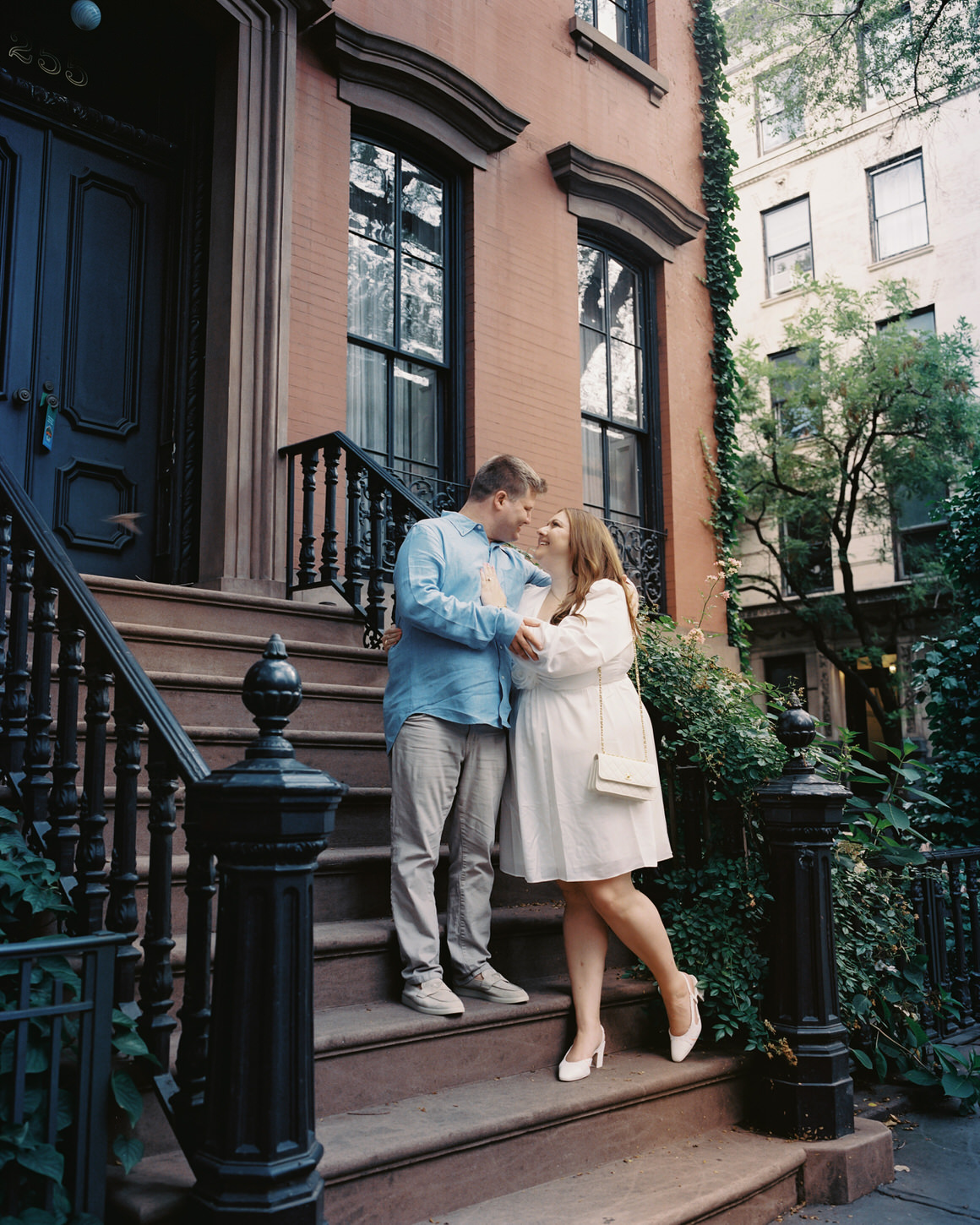 An engaged couple is romantically staring at each other's eyes while standing on a staircase of an ivy-covered building in West Village, NYC, captured by Jenny Fu Studio.