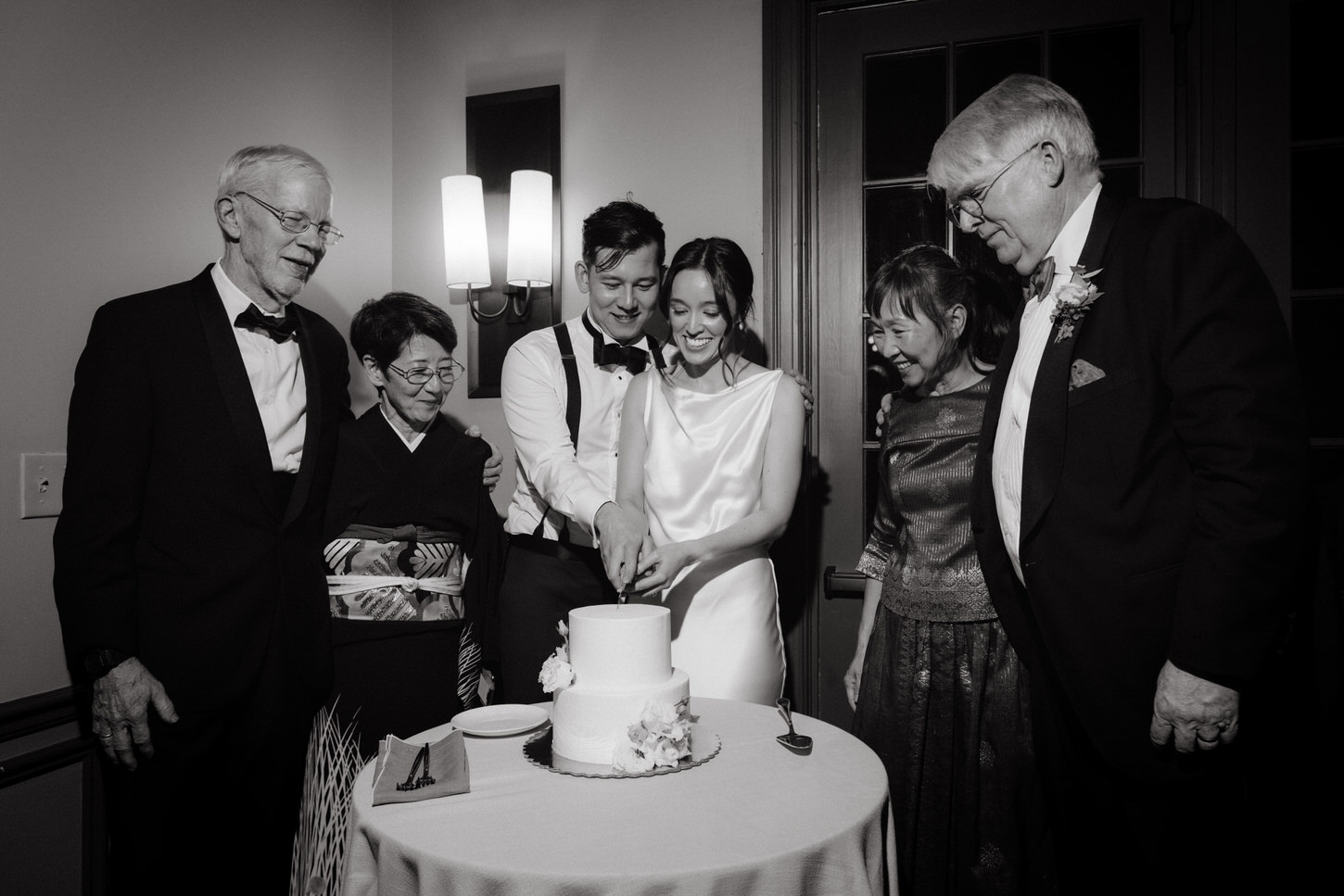 Black and white image of a cake-cutting ceremony with the bride and groom's parents, captured by Jenny Fu Studio.