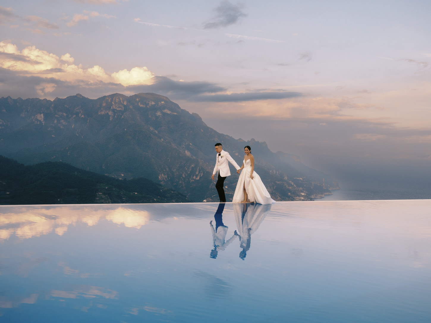 The newlyweds are walking at the edge of an infinity pool, overlooking the grand mountains and lake at Amalfi Coast, Italy, captured by Jenny Fu Studio.