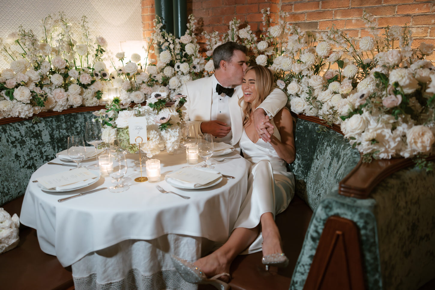 The groom sweetly kisses his bride on the head as they sat in the beautiful wedding reception venue filled with white flowers, captured by Jenny Fu Studio. 