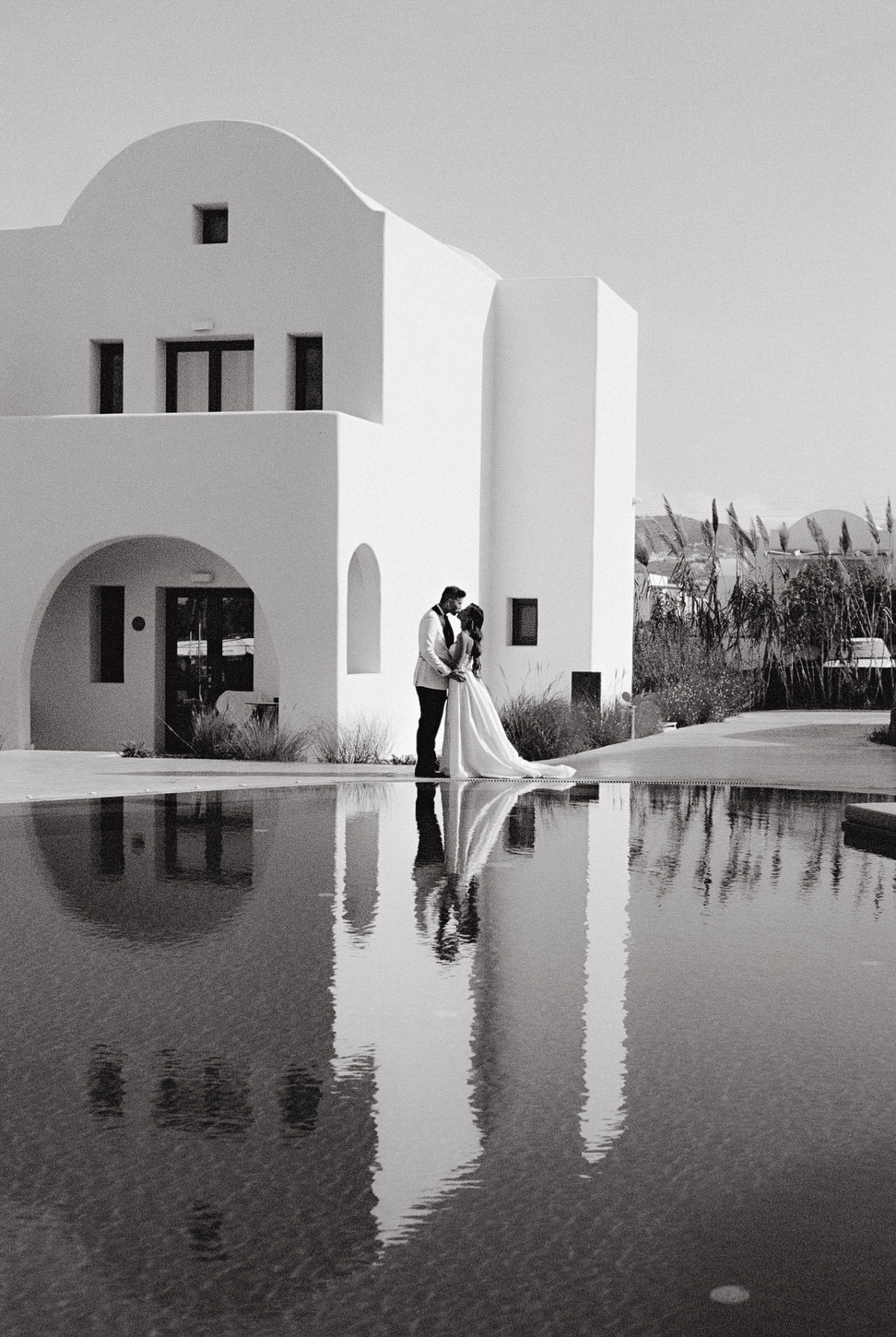 Destination Wedding black and white image of the bride and groom with a villa and pool in the background in Santorini, Greece, captured by Jenny Fu Studio