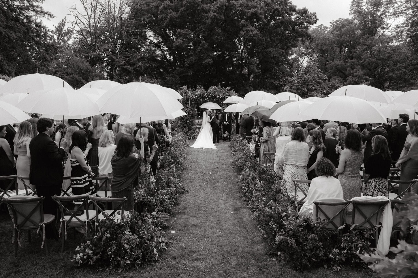 Black and white, timeless image of the wedding ceremony outdoors in the rain, with the guests holding white umbrellas, captured by Jenny Fu Studio.