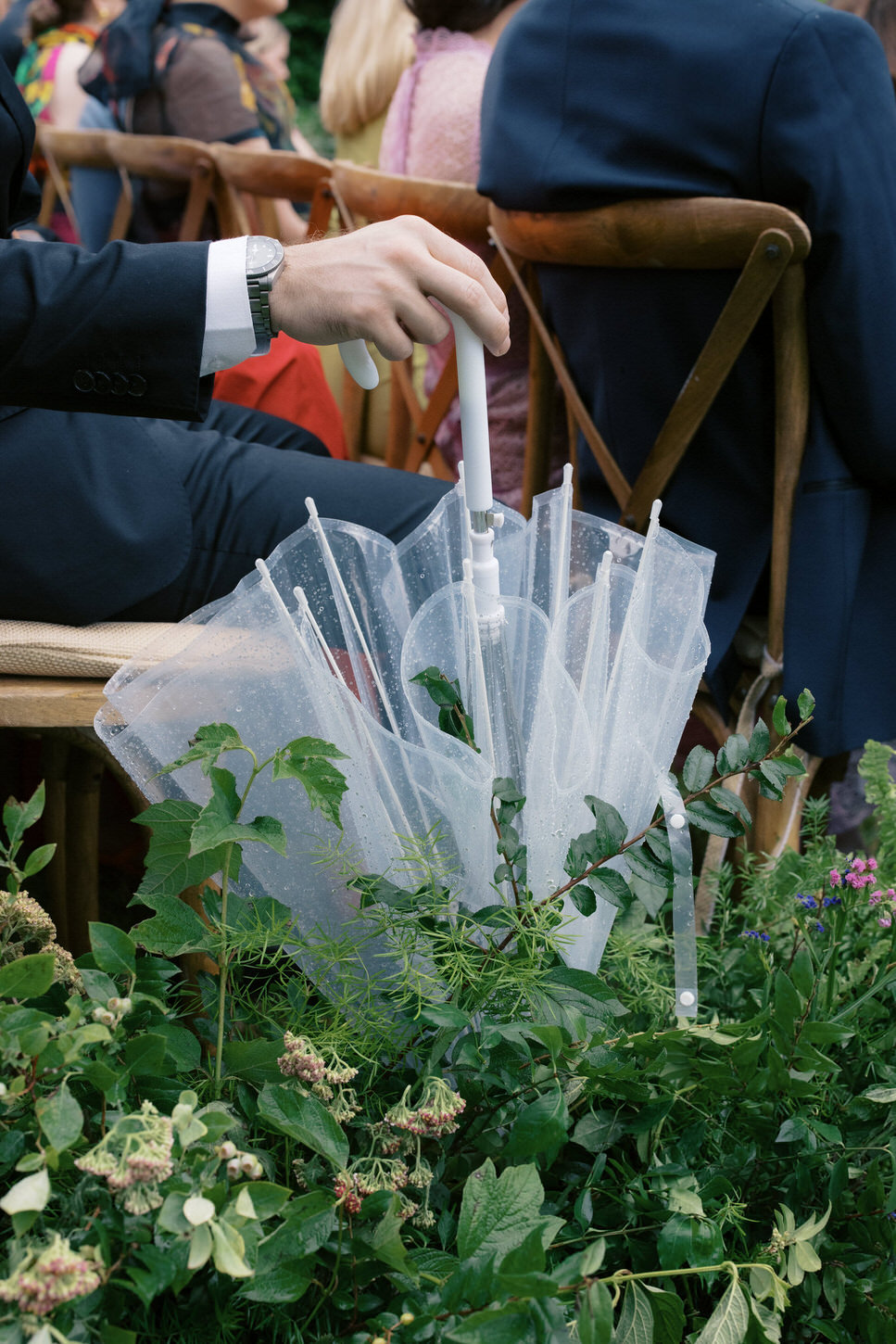 A guest is holding a transparent umbrella while seated at an outdoor wedding ceremony, captured by high-fashion wedding photographer Jenny Fu, of Jenny Fu Studio.