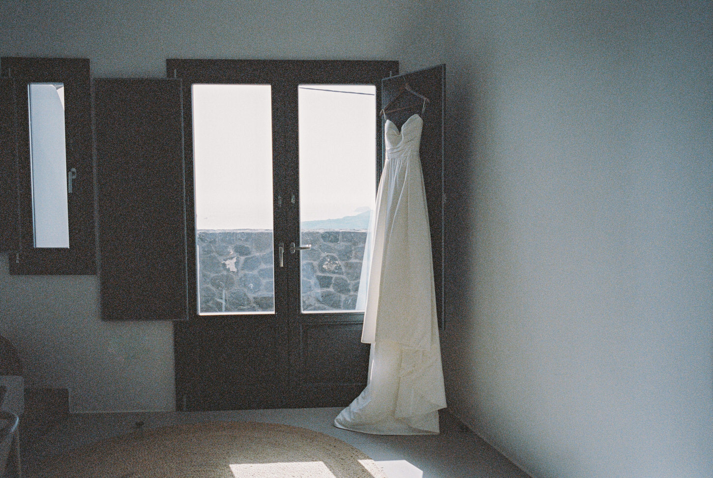 The bride's wedding dress is hanging on a window at Santorini, Greece wedding, captured by Jenny Fu, a Destination Wedding Photographer.