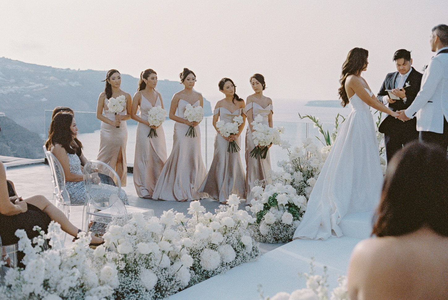 The bridesmaids stand alongside each other, wearing skin-tone dresses in the wedding ceremony at Santorini, Greece, captured by Jenny Fu, a Destination Wedding Photographer.