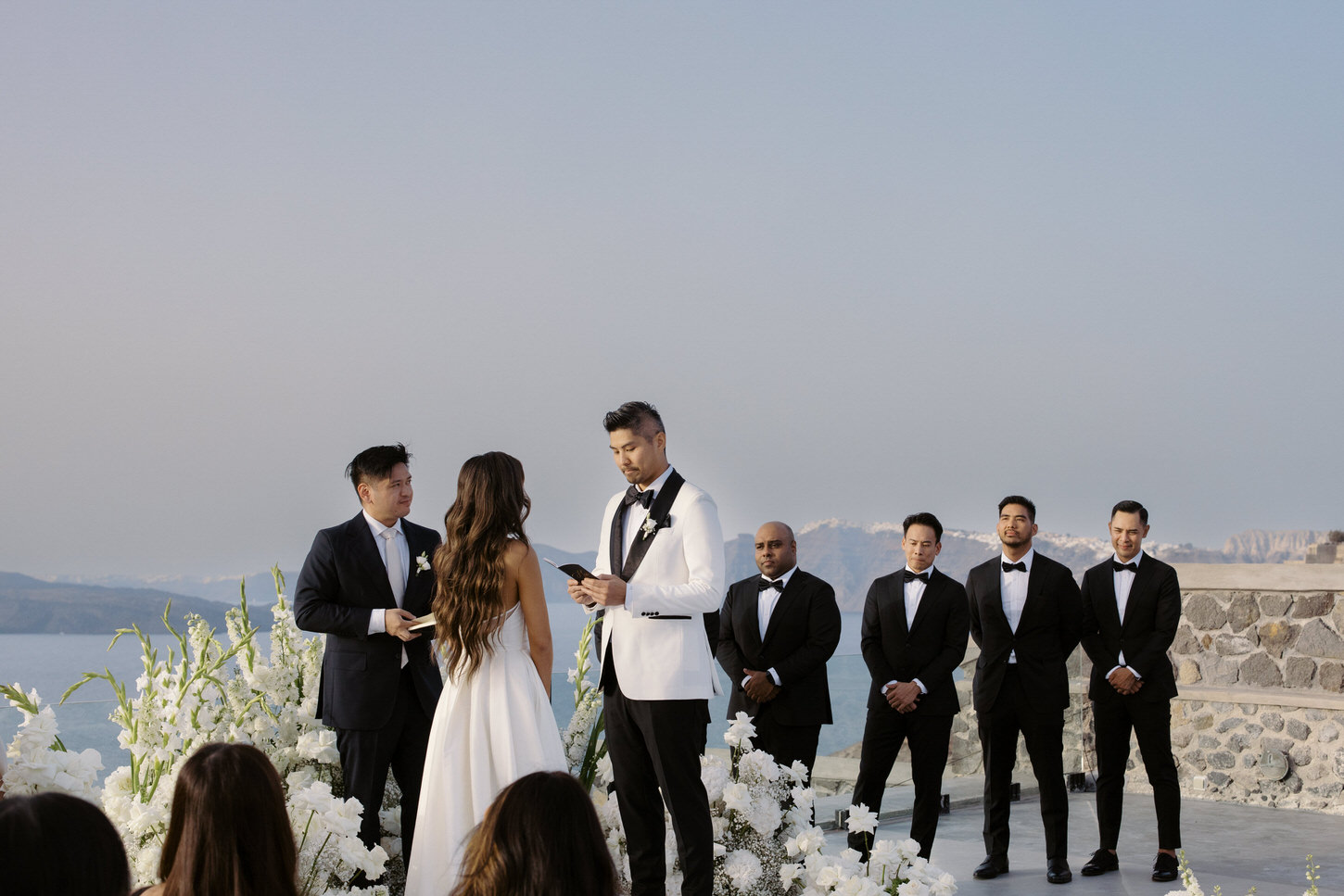 The groomsmen stand alongside each other in the wedding ceremony at Santorini, Greece, captured by Jenny Fu Studio.