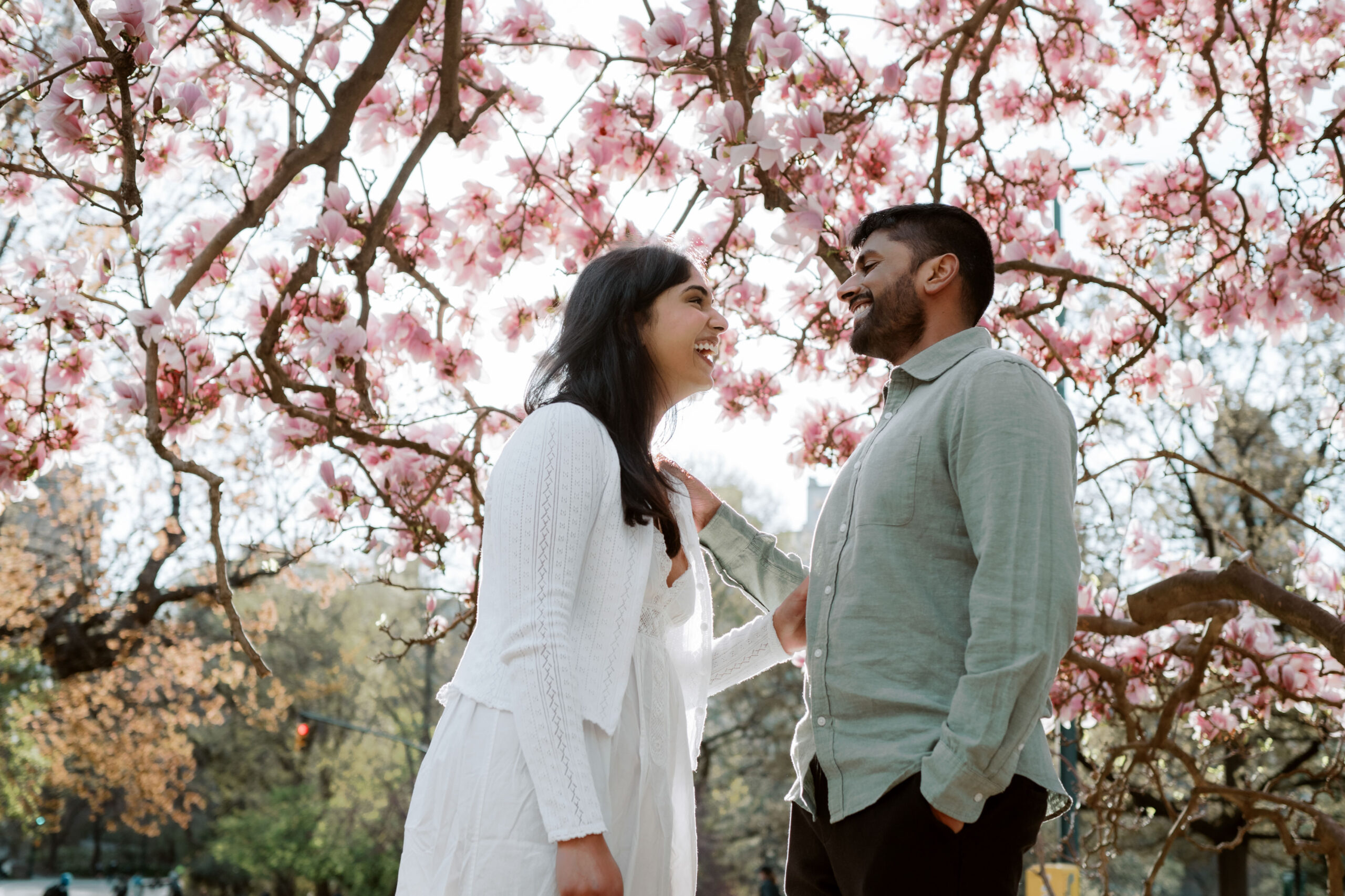 The engaged couple are having a good laugh with cherry blossoms in the background in Central Park, NYC during spring, captured by Jenny Fu Studio for an authentic wedding storytelling.