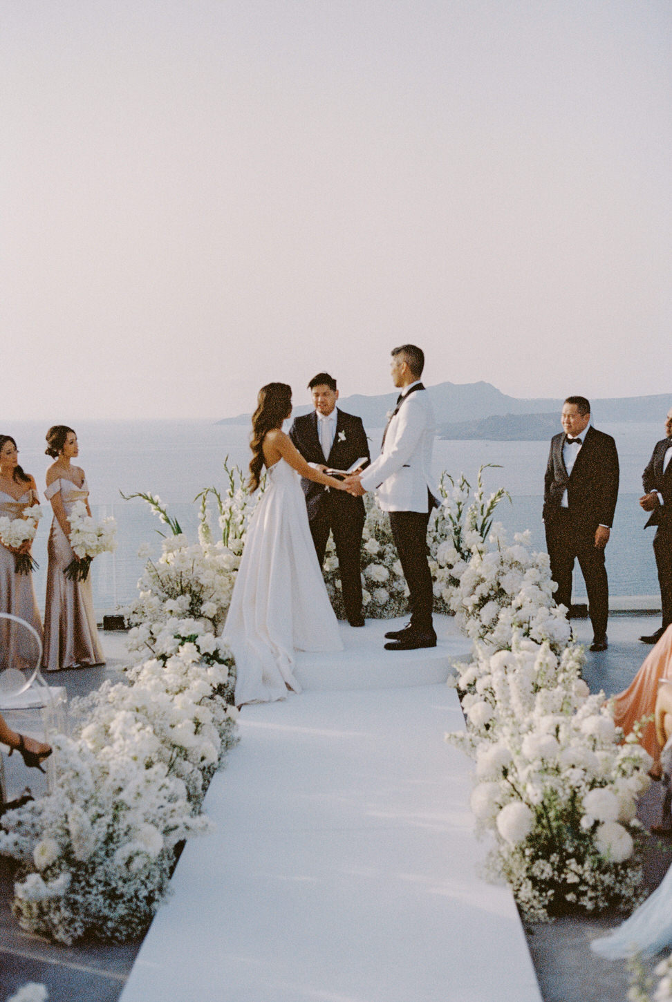 Beautiful wedding ceremony at Santorini, Greece, with white flowers and lake in the background, captured by destination wedding photographer, Jenny Fu Studio.