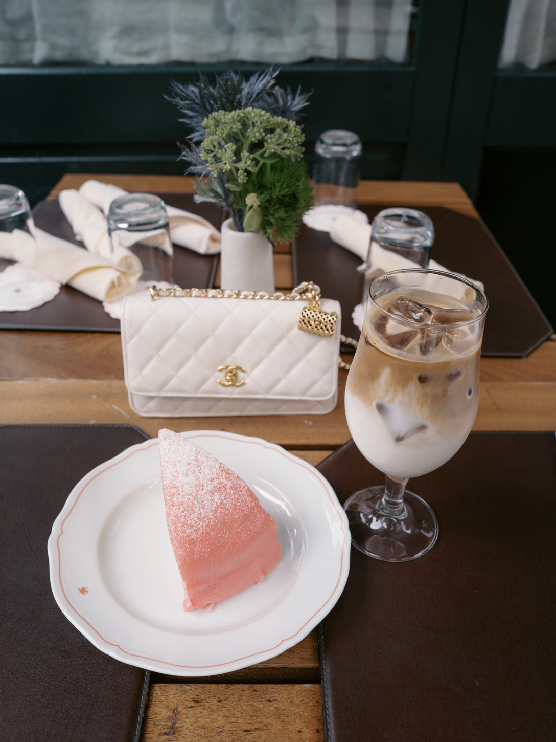 A slice of pink cake, iced coffee drink, and a white Chanel purse on a table at an NYC restaurant where the engaged couple went, captured by Jenny Fu Studio.