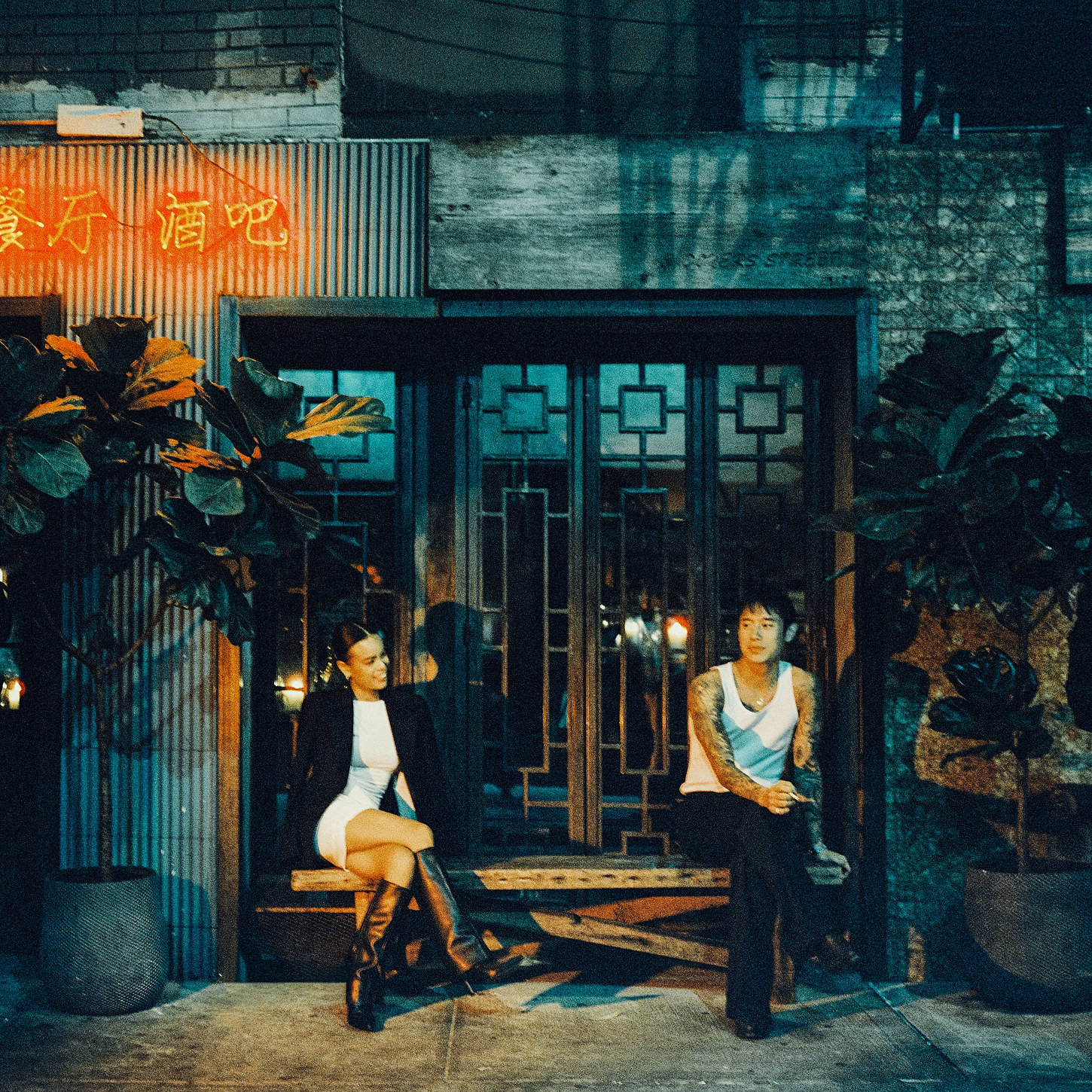 Couple sitting down, in front of a neon-lit Chinatown shop, having a chill night during their nighttime wedding shoot.