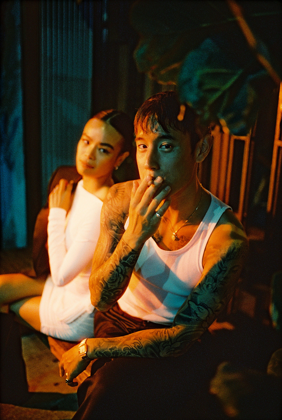 Groom with tattoos smokes under neon light, bride blurred in background, Chinatown elopement photo.