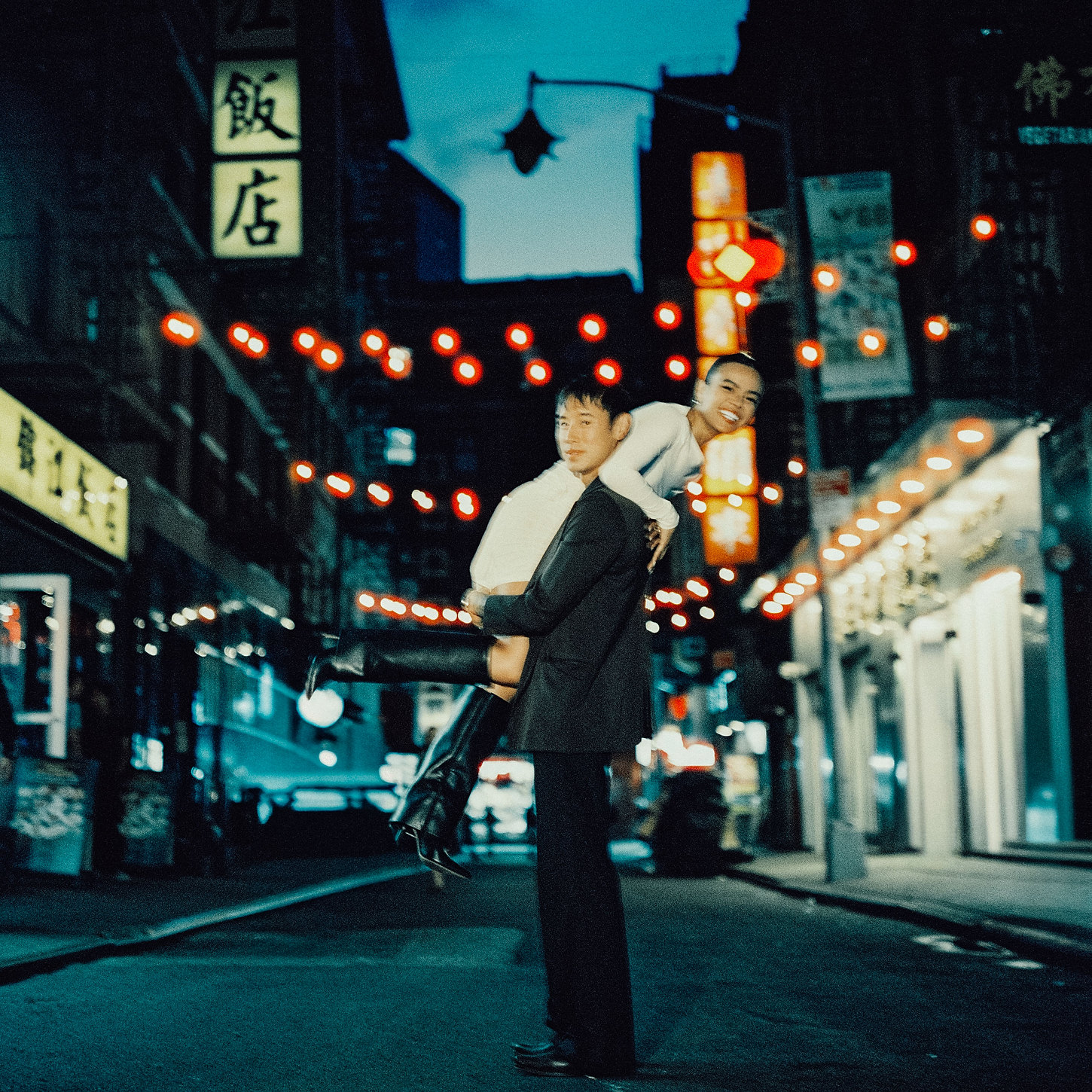 The groom carries his happy bride under Chinatown string lights during a film wedding photography shoot.