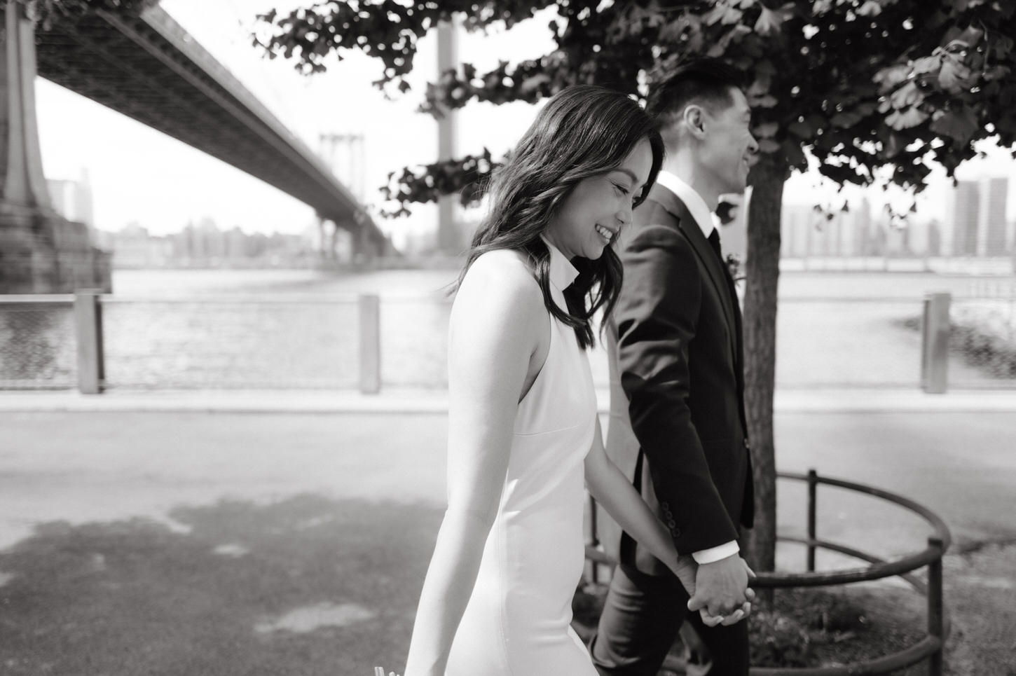 The newlyweds walks hand in hand, happily, with the Brooklyn Bridge in the background, in a documentary-style wedding photo by Jenny Fu Studio, NYC.